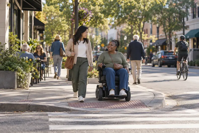 People crossing a sunny main street, one using a power wheelchair